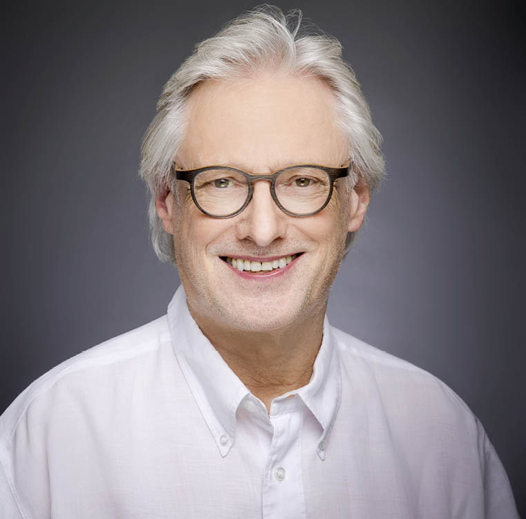 Eine l chelnde Person mit grauem Haar, Brille und wei em Hemd vor wei em Hintergrund. Beschreibung von KI generiert