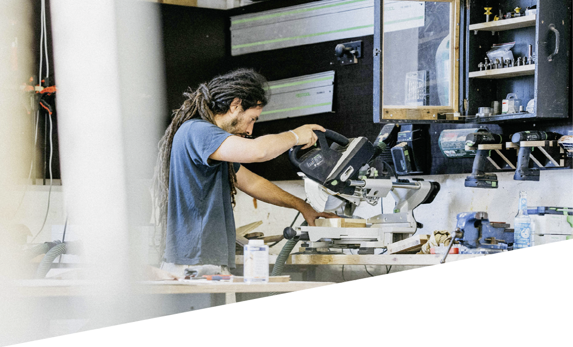 Eine Person mit langen Dreadlocks und blauem T-Shirt arbeitet konzentriert an einer Werkbank mit einer elektrischen S ge in einer Werkstatt. Beschreibung von KI generiert
