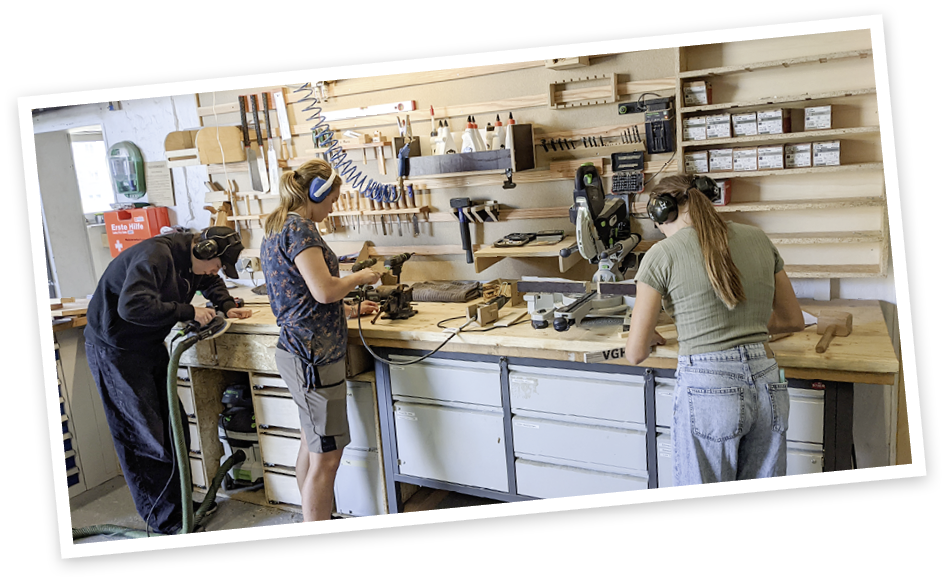 Drei Personen mit Geh rschutz arbeiten konzentriert an einer Werkbank in einer Holzwerkstatt, umgeben von Werkzeugen und Materialien. Beschreibung von KI generiert