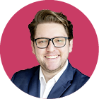Eine l chelnde Person mit kurzen, zur ckgek mmten Haaren tr gt eine Brille und einen dunklen Anzug vor einem roten Hintergrund. Beschreibung von KI generiert