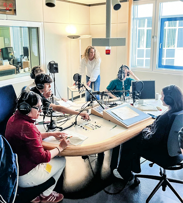 Mehrere Personen mit Kopfh rern sitzen an einem Tisch mit Mikrofonen in einem hellen Radiostudio, w hrend eine weitere Person im Hintergrund steht. Beschreibung von KI generiert