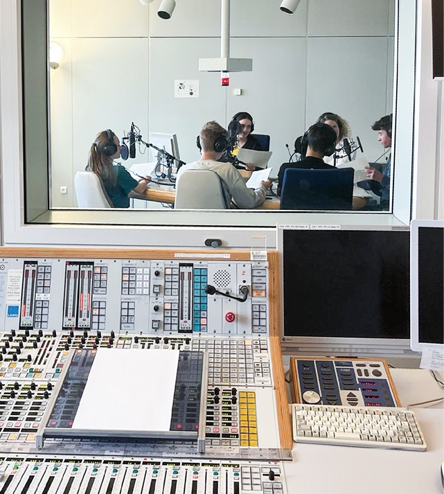 In einem Tonstudio sitzen mehrere Personen mit Kopfh rern an Mikrofonen und sprechen, w hrend im Vordergrund ein Mischpult und zwei Bildschirme zu sehen sind. Beschreibung von KI generiert