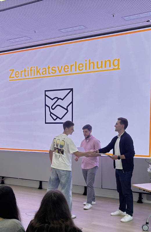 Sechs Personen sitzen an einem Tisch in einem modernen Besprechungsraum und arbeiten konzentriert an Unterlagen, w hrend auf einem Bildschirm eine Pr sentation mit farbigen K stchen und Daumen-hoch- und Daumen-runter-Symbolen angezeigt wird. Beschreibung von KI generiert