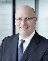 Eine Person mit Brille tr gt einen dunklen Anzug, ein wei es Hemd und eine gemusterte Krawatte und l chelt vor einem unscharfen B rohintergrund. Beschreibung von KI generiert