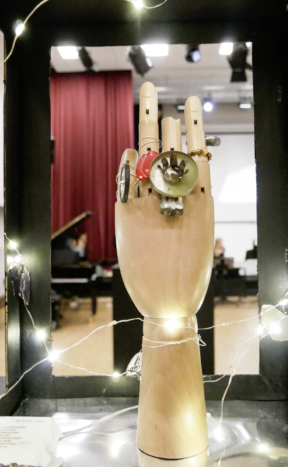 Eine h lzerne Hand mit mehreren auff lligen Ringen steht aufrecht in einem beleuchteten Rahmen vor einem Raum mit rotem Vorhang und Klavier. Beschreibung von KI generiert
