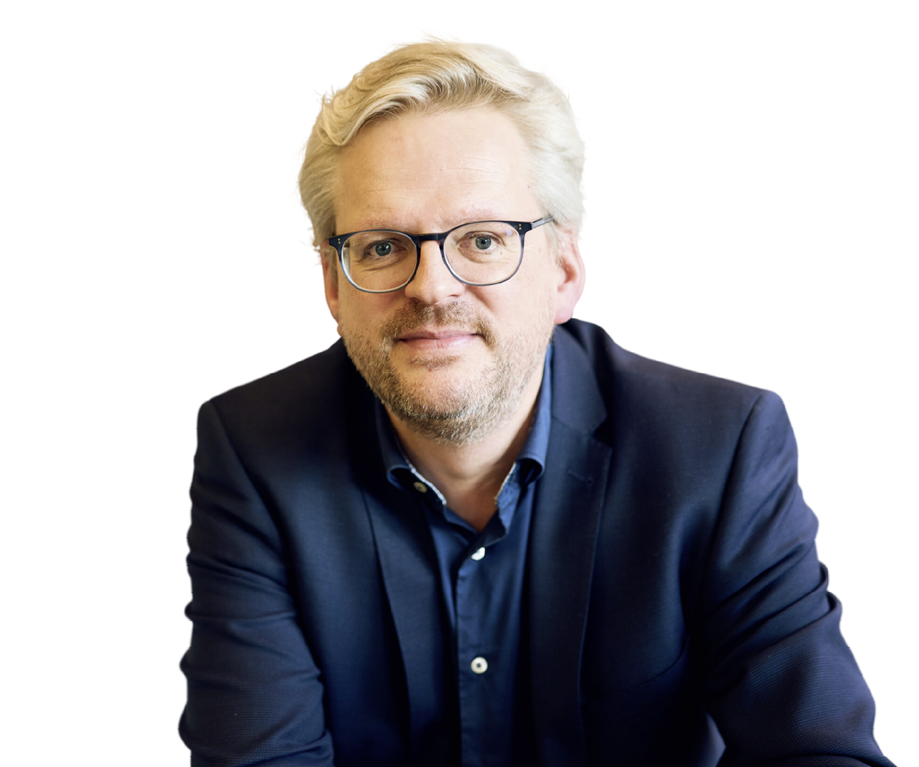 Eine Person mit hellen, zur ckgek mmten Haaren tr gt einen dunklen Anzug und ein dunkles Hemd und sitzt vor einem wei en Hintergrund. Beschreibung von KI generiert