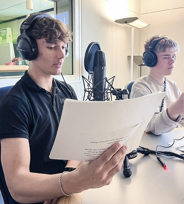 Zwei Personen mit Kopfh rern sitzen an einem Tisch vor einem Mikrofon, wobei eine Person ein Blatt Papier h lt und zu sprechen scheint. Beschreibung von KI generiert