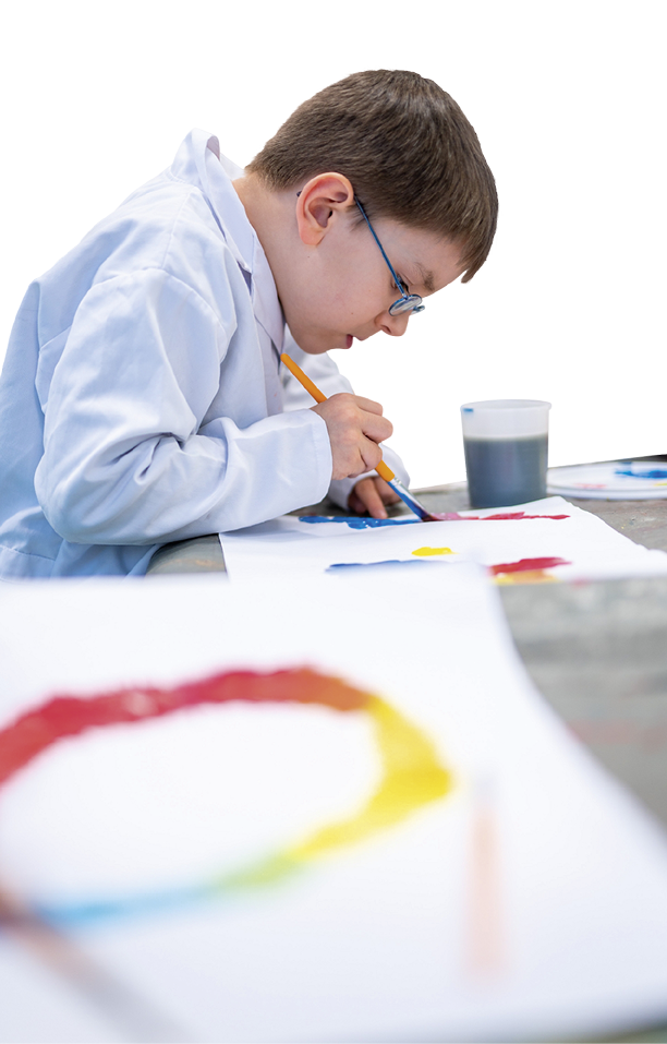 Ein Kind mit Brille und wei em Kittel sitzt an einem Tisch und malt konzentriert mit einem Pinsel auf Papier. Beschreibung von KI generiert