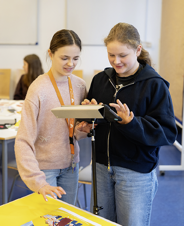 Zwei Personen stehen in einem Klassenraum und bedienen gemeinsam ein Ger t auf einem Stativ, w hrend sie auf einen Tisch mit einem bunten Bild schauen. Beschreibung von KI generiert