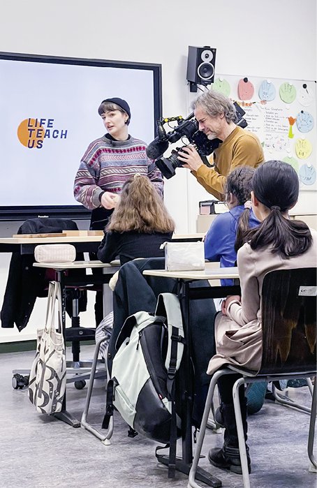 In einem Klassenzimmer steht eine Person mit einem bunten Pullover vor einer Gruppe sitzender Personen, w hrend eine weitere Person mit einer Kamera filmt und im Hintergrund ein Bildschirm mit der Aufschrift „LIFE TEACH US“ zu sehen ist. Beschreibung von KI generiert
