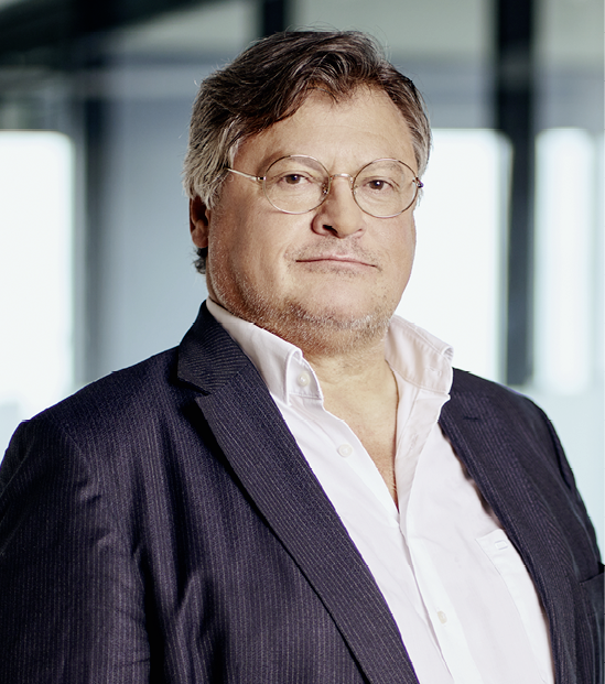 Eine Person mit Brille tr gt ein wei es Hemd und einen dunklen Anzug und steht vor einem unscharfen, modernen Hintergrund. Beschreibung von KI generiert
