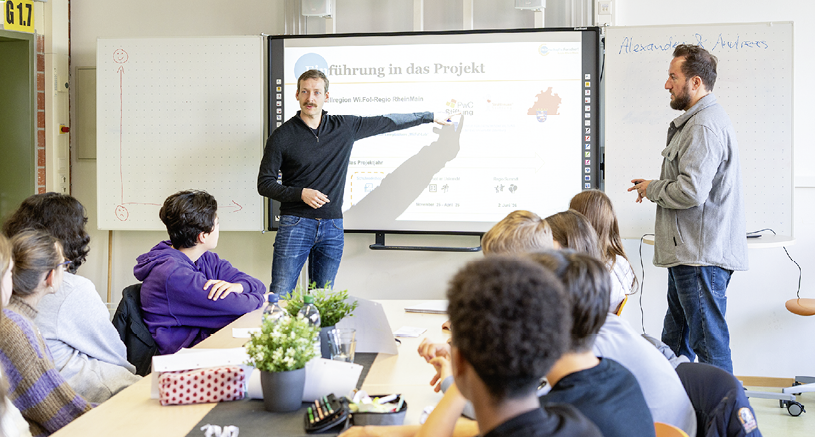 Mehrere Personen sitzen an einem Tisch und h ren zwei Erwachsenen zu, die vor einem Bildschirm mit einer Pr sentation in einem Klassenraum stehen. Beschreibung von KI generiert
