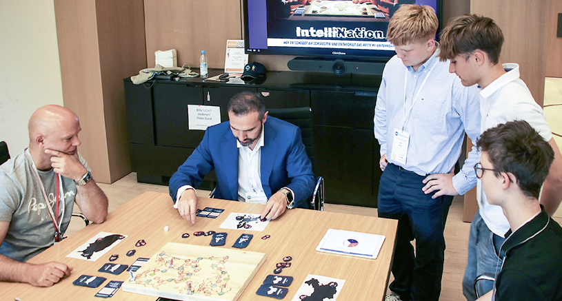 F nf Personen mit Namensschildern spielen und beobachten ein Brettspiel an einem Tisch in einem modernen Innenraum mit einem Bildschirm im Hintergrund. Beschreibung von KI generiert
